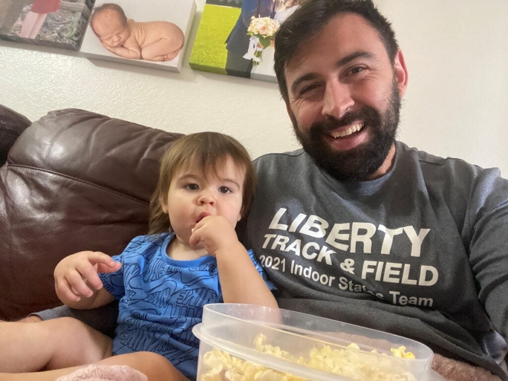 A man with a dark beard takes a selfie sitting on his couch while eating popcorn with his young son.