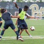 Four people playing soccer together on a soccer field. They are each wearing shorts and t-shirts, with two wearing neon vests over their shirts.