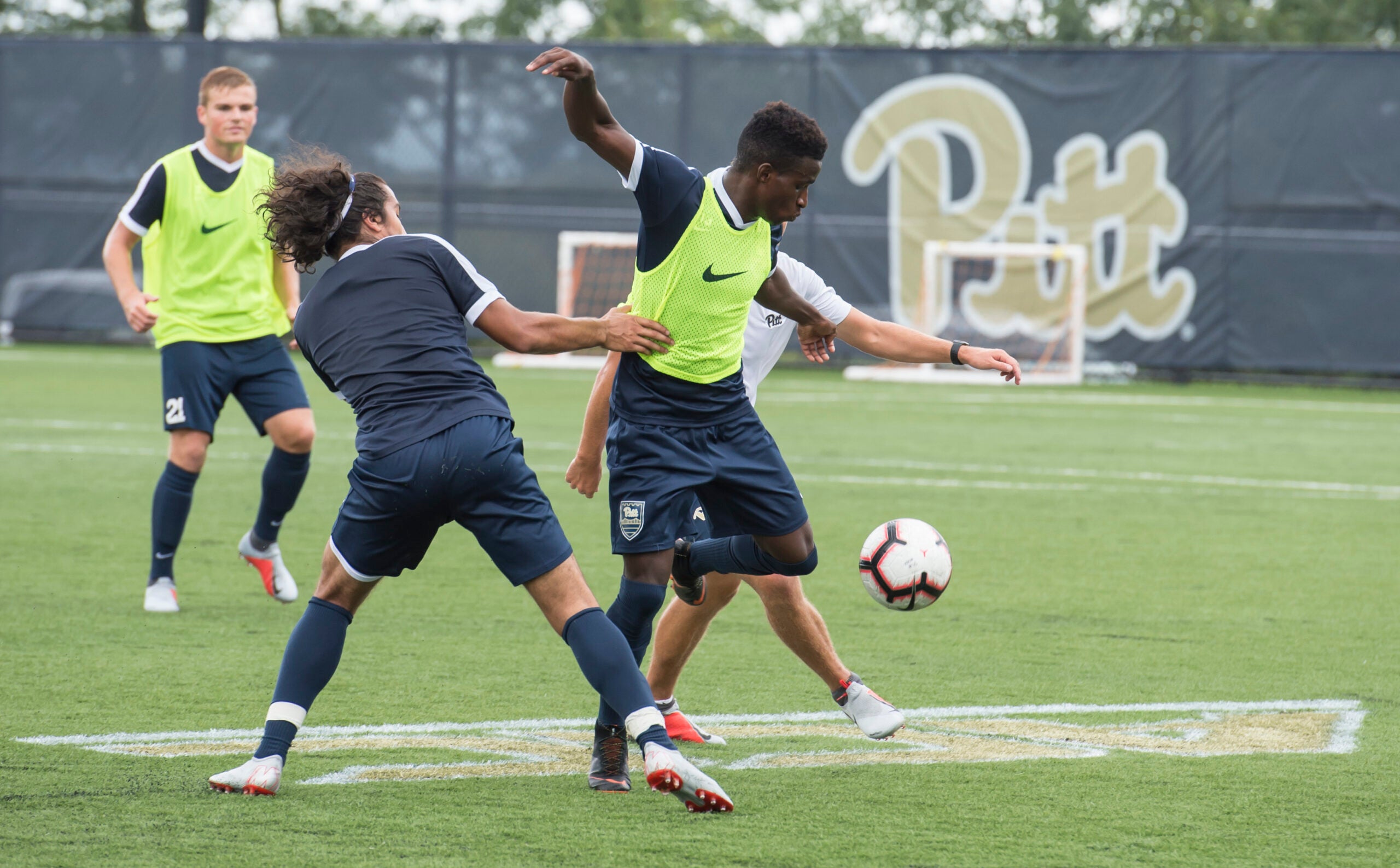 Four people playing soccer together on a soccer field. They are each wearing shorts and t-shirts, with two wearing neon vests over their shirts.