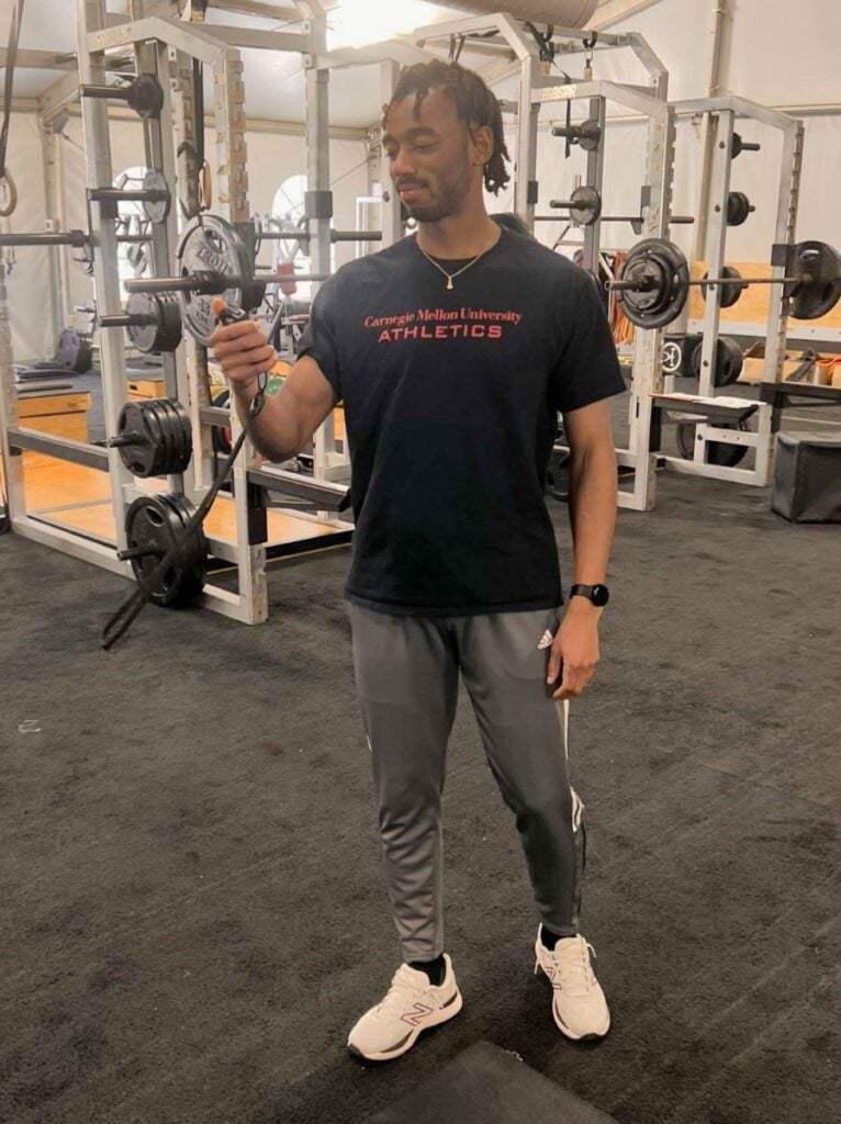 A man with medium skin tone and short black hair wearing gray athletic pants and a Carnegie Mellon University Athletics t-shirt holds a device while standing in a gym