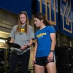 Two women wearing Pitt Athletics clothing look at a tablet device while standing in a gym