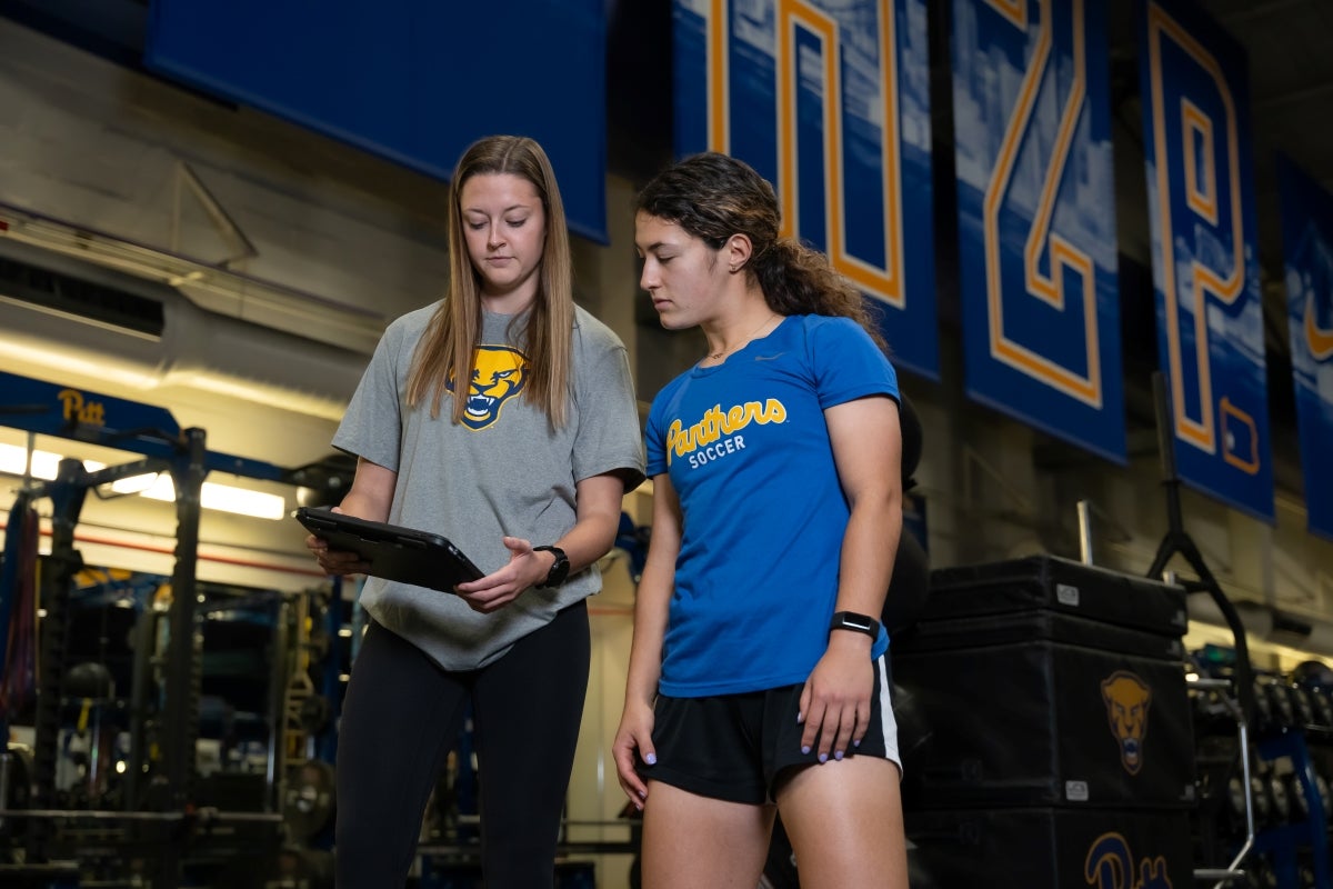 Two women wearing Pitt Athletics clothing look at a tablet device while standing in a gym