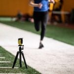 A monitoring device sits on a small tripod at the edge of an indoor field while a woman in black tights and blue shirt runs next to it