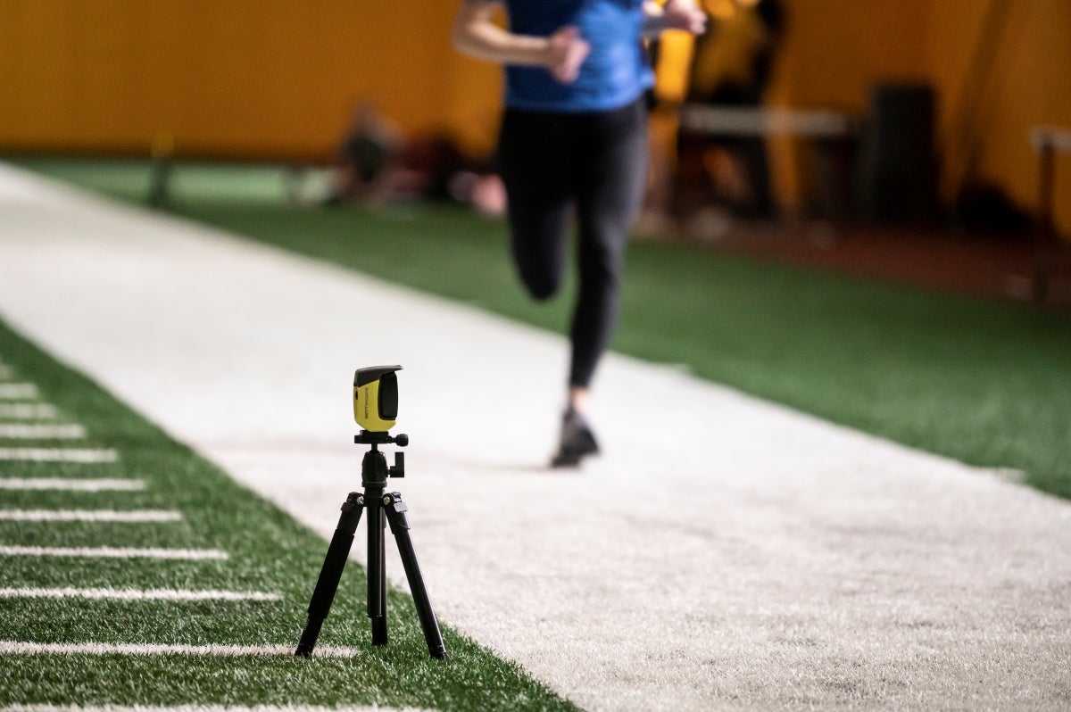 A monitoring device sits on a small tripod at the edge of an indoor field while a woman in black tights and blue shirt runs next to it