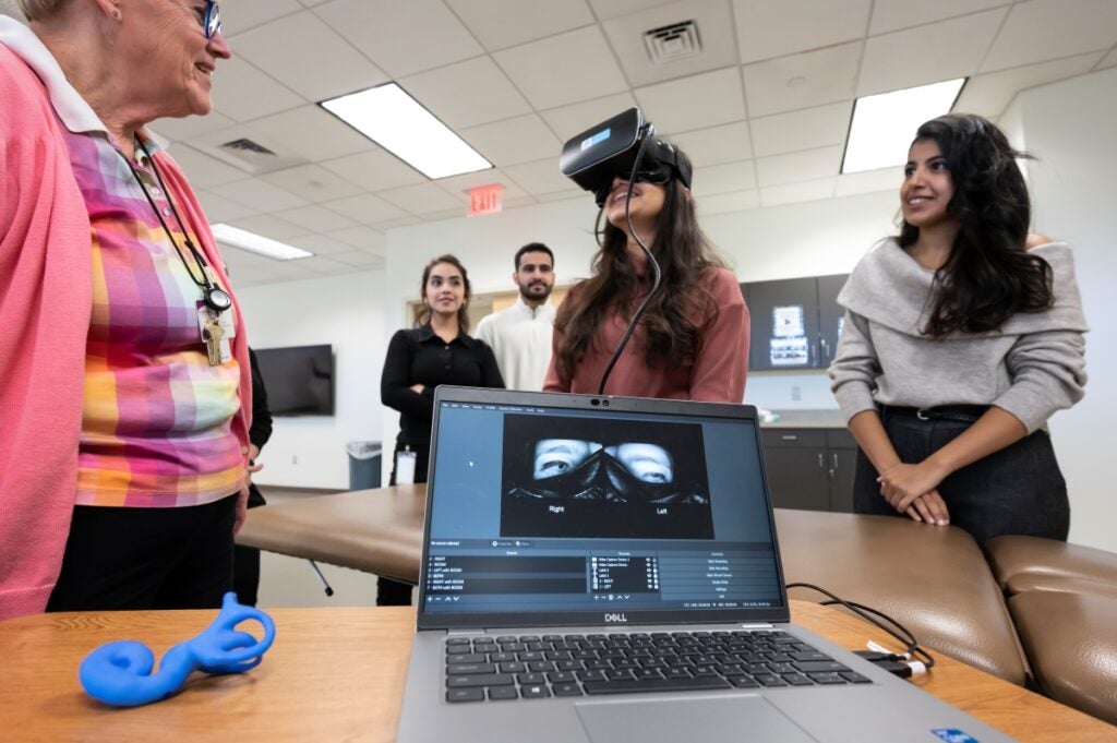 A student is standing and wearing specialized headgear covering her eyes with a long cord connected to a computer. A professor and other students look on. The laptop computer screen shows software with an image of the student's eyes.