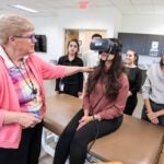 A female professor wearing a pink sweater instructs a student wearing large, specialized goggles while classmates look on