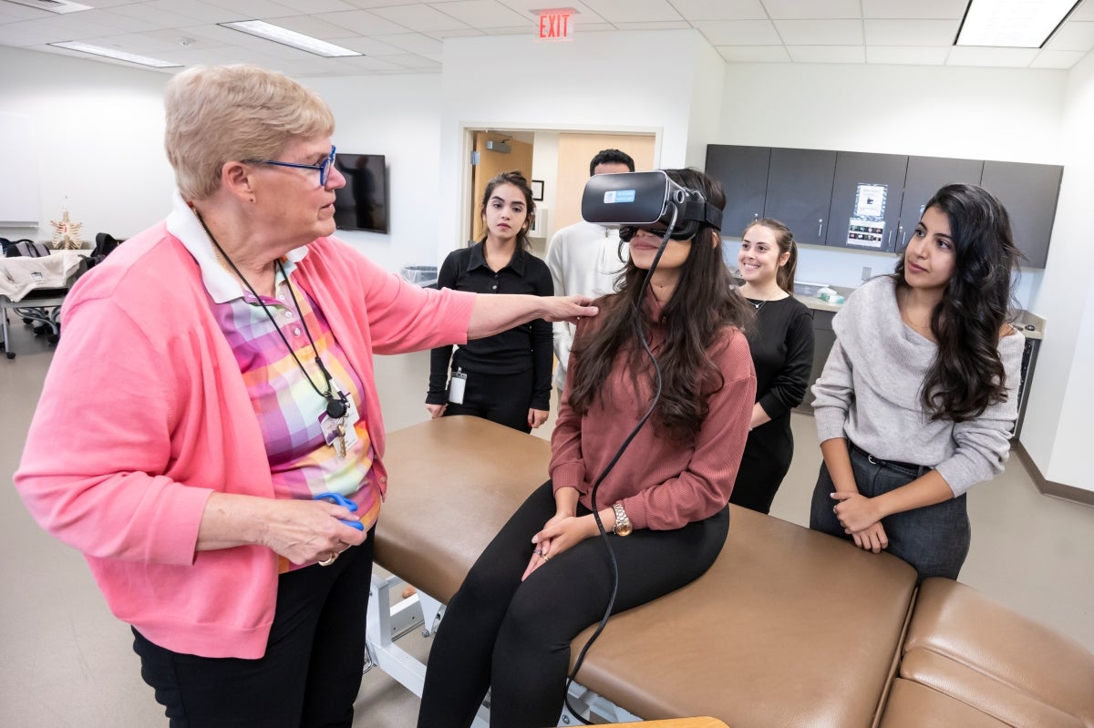 A female professor wearing a pink sweater instructs a student wearing large, specialized goggles while classmates look on