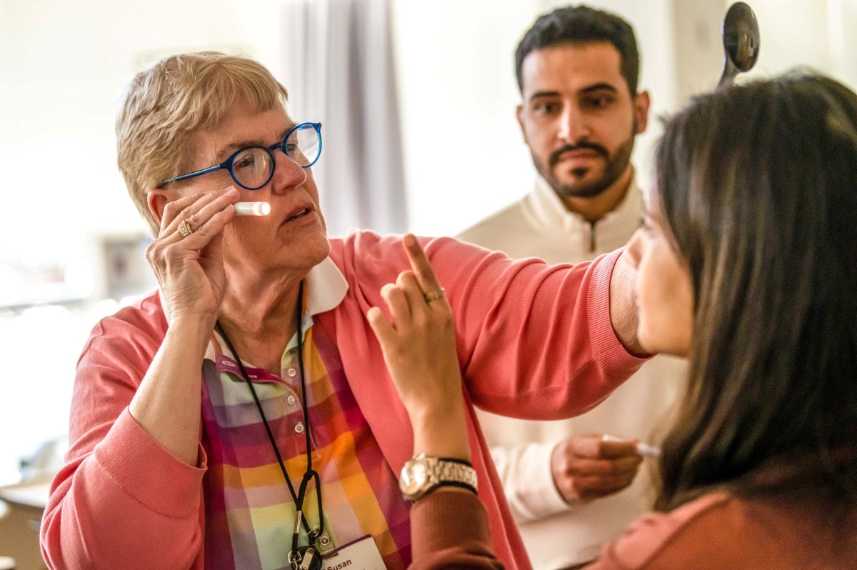 A woman with short blonde hair wearing a peach cardigan over a red, pink and white collared shirt and blue glasses holding a flashlight examining a woman with brown hair wearing a red shirt while a man with dark hair wearing a white shirt standings in the background.