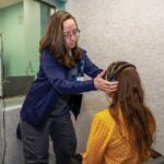 A woman with long brown hair and glasses adjusts headphones on a woman sitting in a chair