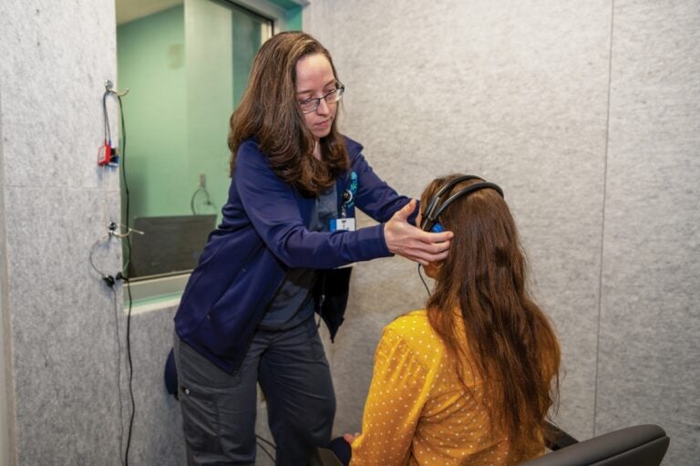 A woman with long brown hair and glasses adjusts headphones on a woman sitting in a chair