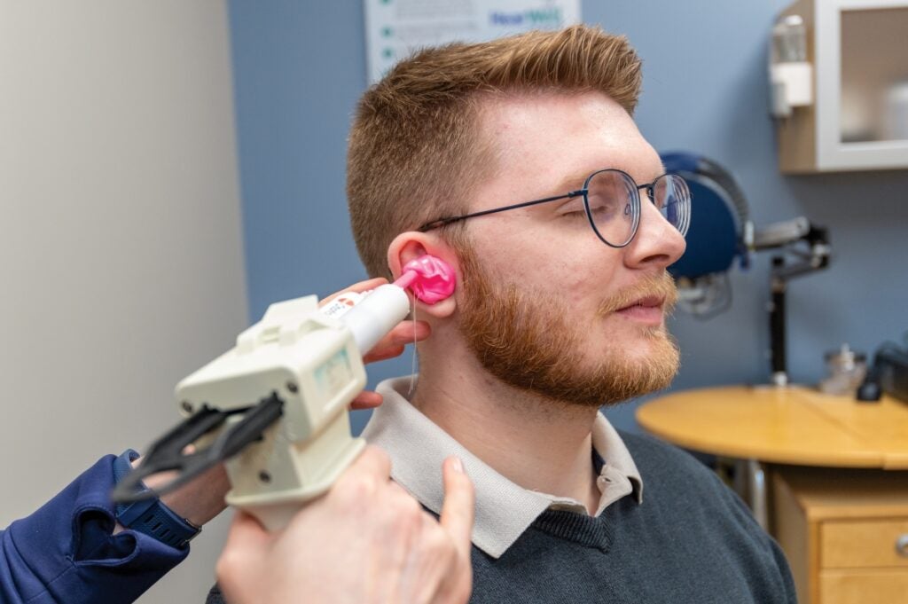 A man with short red hair and a beard wearing glasses has a hand held testing device held up to his right ear by a technician