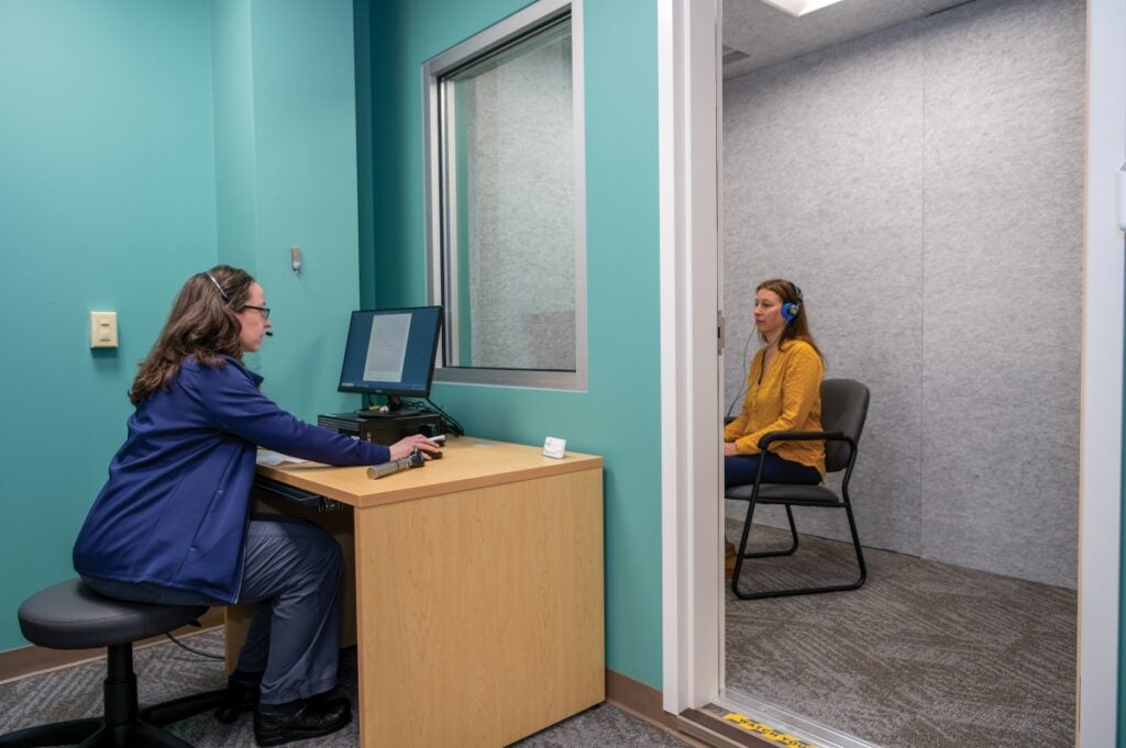 A woman in a blue lab coat wearing headphones sits at a computer while on the other side of a glass window a woman sits in a chair wearing headphones