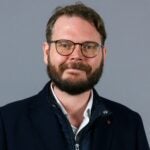 Man with brown hair and brown beard wearing glasses and a dark blue jacket