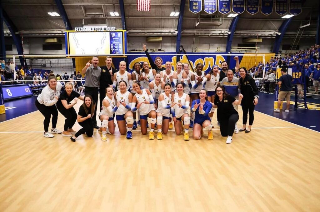 A group of 26 women's volleyball players and staff stand and kneel on the volleyball court while smiling and raising their arms in celebration