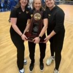 Three women stand together smiling as their arms hold a large rectangular trophy etched with a volleyball and NCAA.