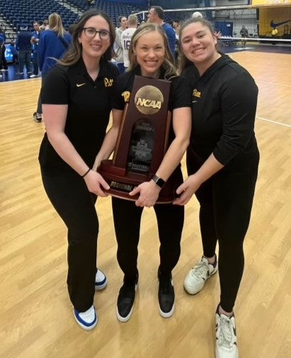 Three women stand together smiling as their arms hold a large rectangular trophy etched with a volleyball and NCAA.