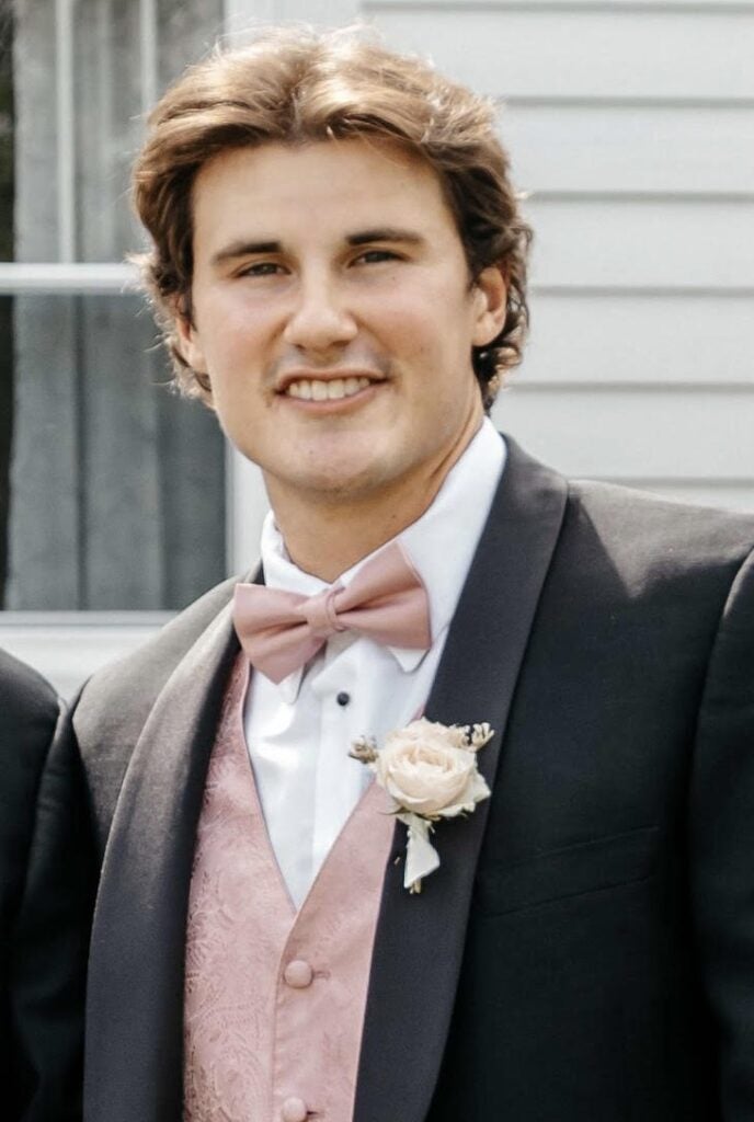 A man with short brown hair smiles at the camera while wearing a formal black suit