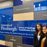 Two women with long black hair stand in front of a wall with names of the SHRS disciplines