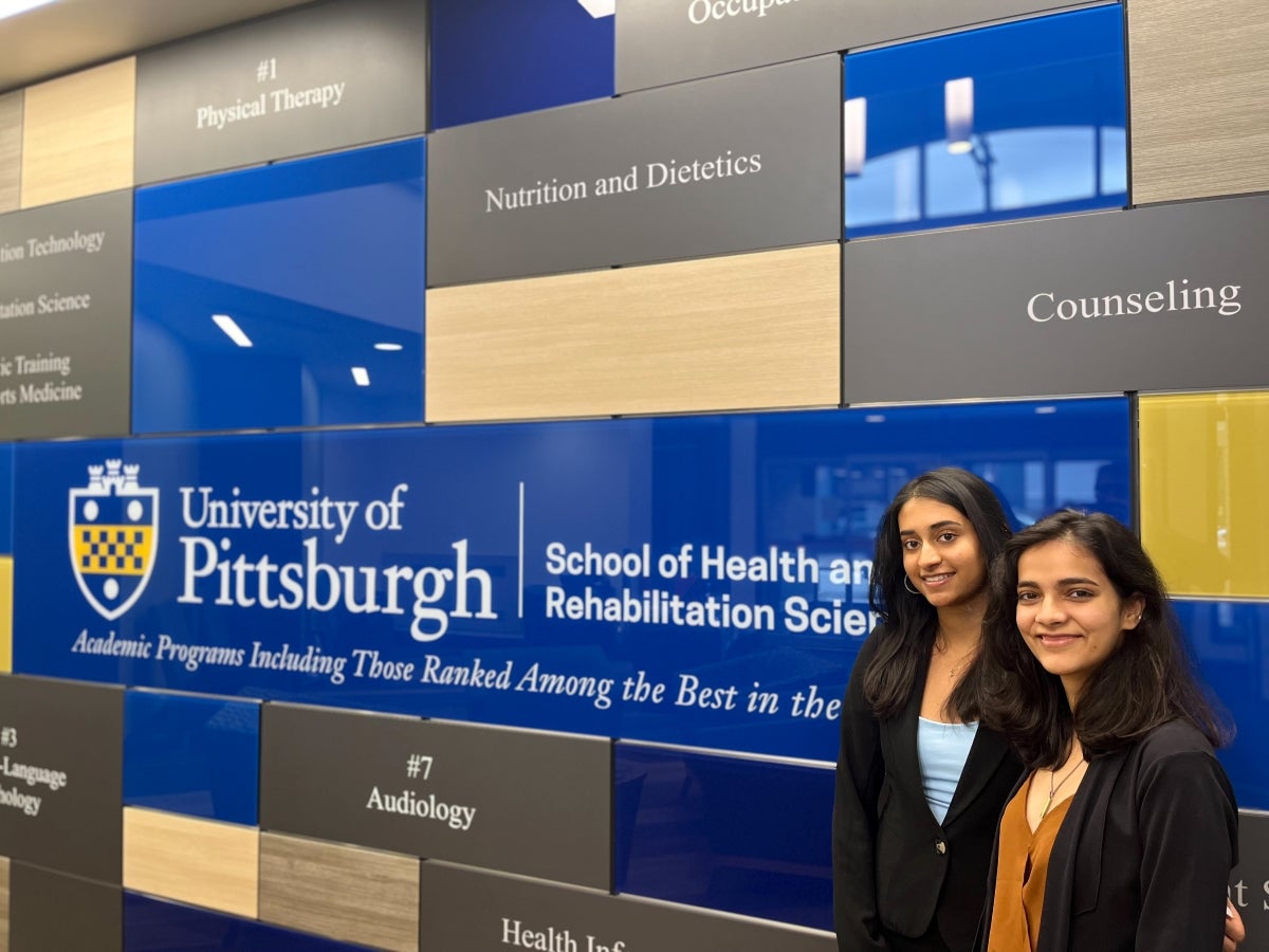 Two women with long black hair stand in front of a wall with names of the SHRS disciplines