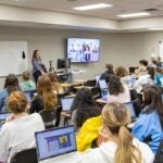 A classroom full of students sitting at desks with laptops while a teacher stands at the front of the room speaking to the class.