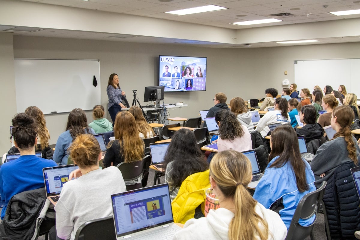 A classroom full of students sitting at desks with laptops while a teacher stands at the front of the room speaking to the class.