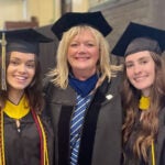 Three women standing together wearing black graduation gowns and caps and blue and gold sashes.