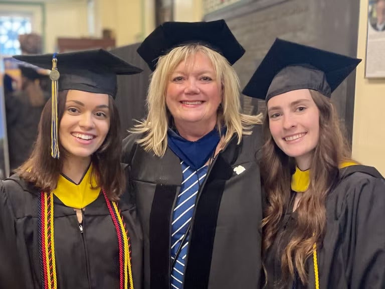 Three women standing together wearing black graduation gowns and caps and blue and gold sashes.