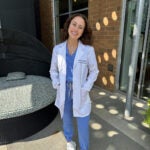 Woman with brown hair wearing blue scrubs, white sneakers and a white lab coat.