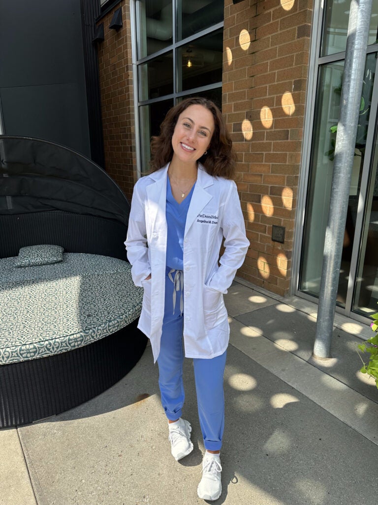 Woman with brown hair wearing blue scrubs, white sneakers and a white lab coat.