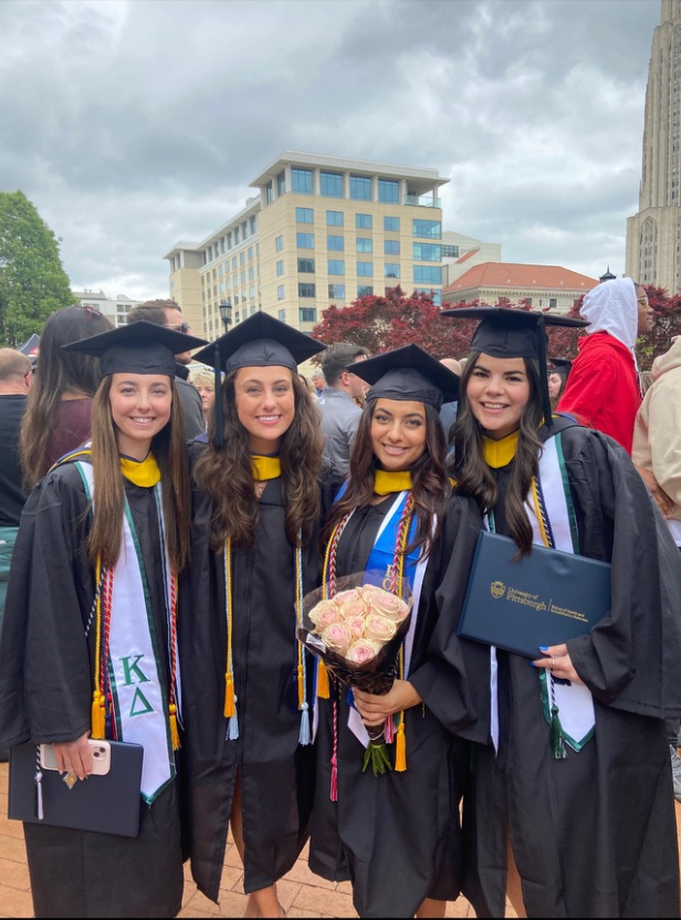 Four women with brown hair wearing graduation caps and gowns wearing academic cords and holding flowers and diplomas.