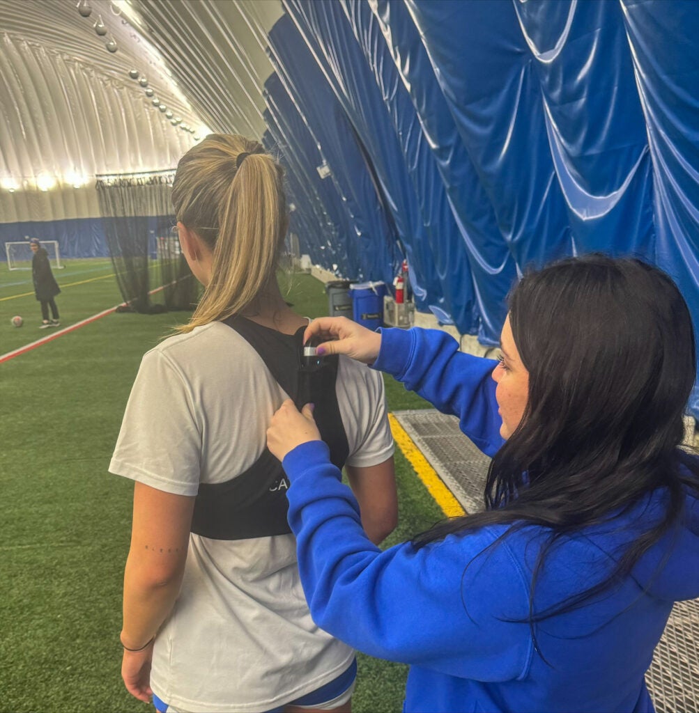 Brunette woman (right) wearing a blue hoodie placing a sports science device on a blonde woman in front of her who is wearing a tan shirt.