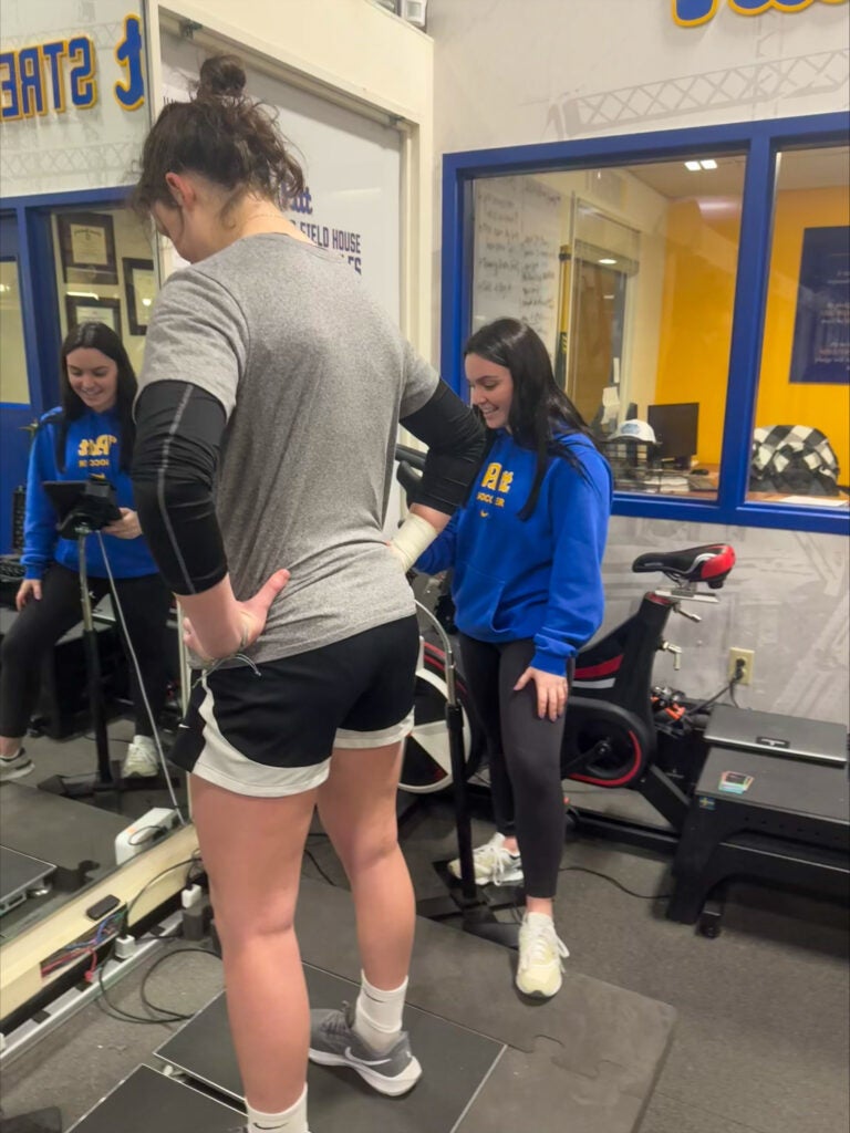 A woman with brown hair in a bun wearing a gray t shirt over black long sleeves and black and white shorts, standing on a platform next to a woman with brown hair wearing a blue Pitt hoodie and black leggings.