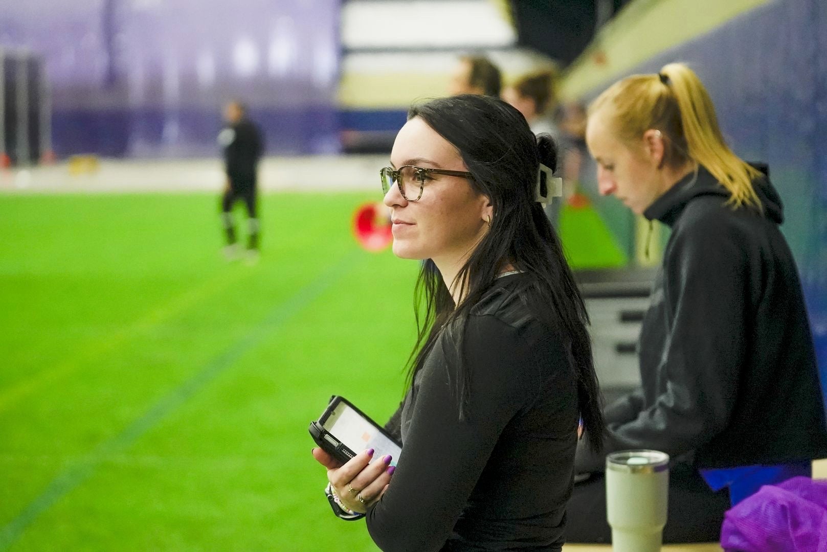 Woman with dark brown hair clipped in the back wearing glasses and a black long sleeve shirt holding a tablet while looking onward.