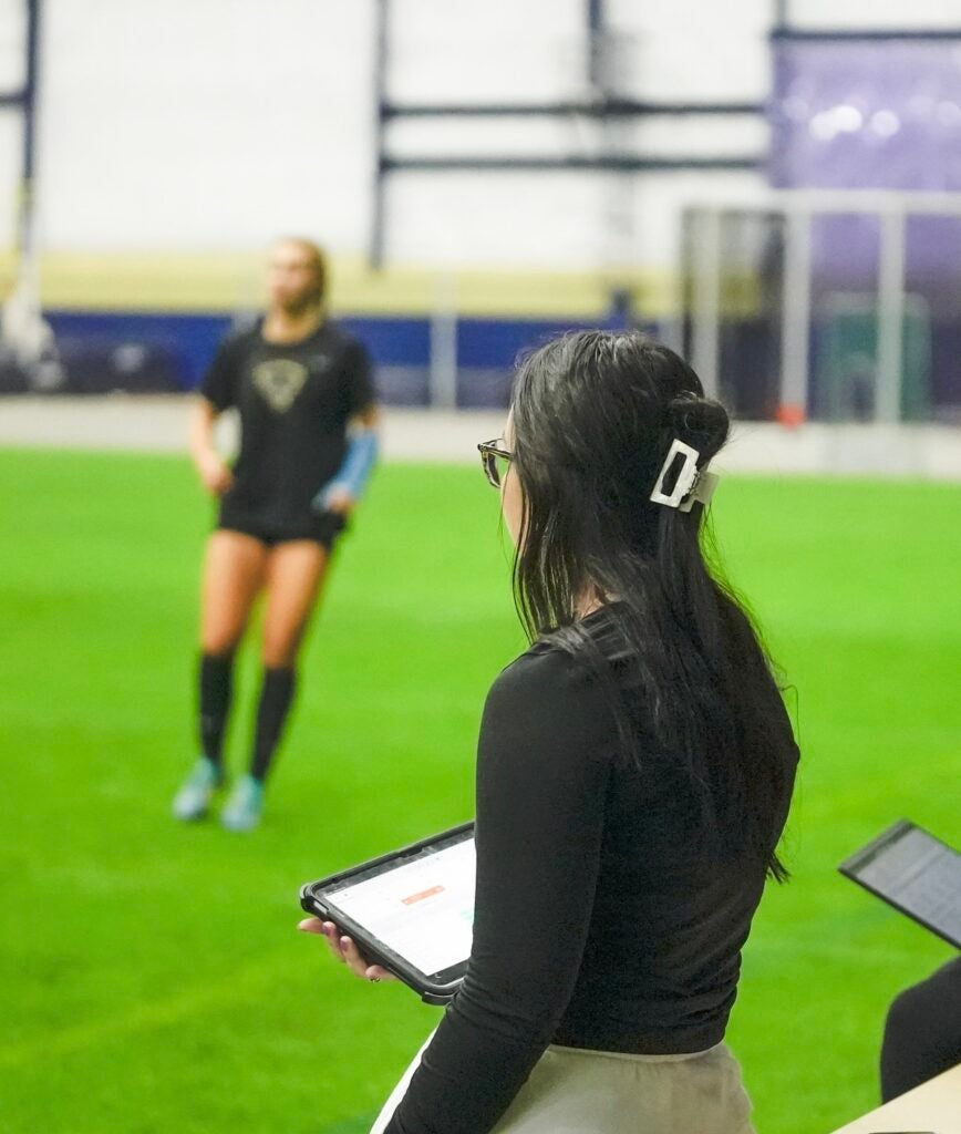 Woman with dark brown hair with her hair clipped back wearing a long sleeve black shirt and white pants while holding a tablet looks to a female athlete who is out of focus.