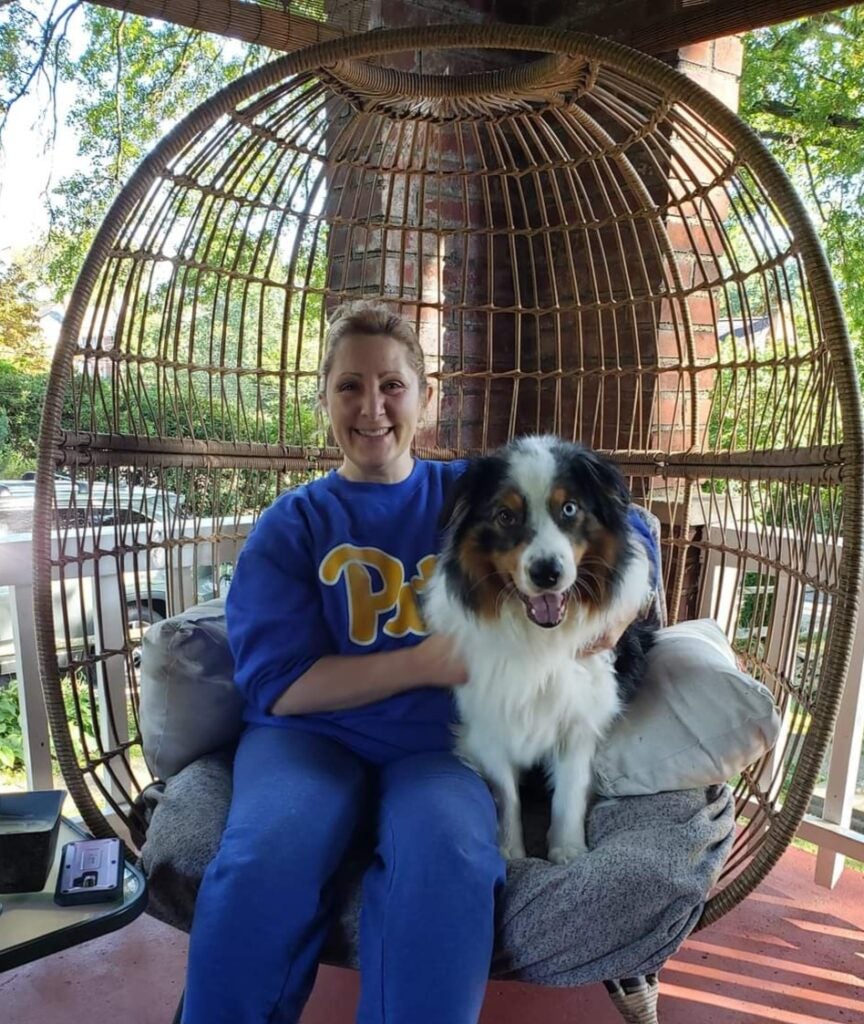 A woman with her hair pulled back wearing a royal blue sweatsuit sitting with an Australian Shepherd on a porch.