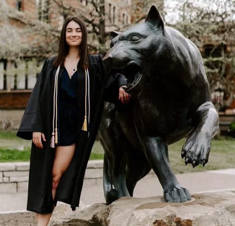 A woman with brown hair wearing a black graduation gown over a black dress standing next to a statue of a panther.