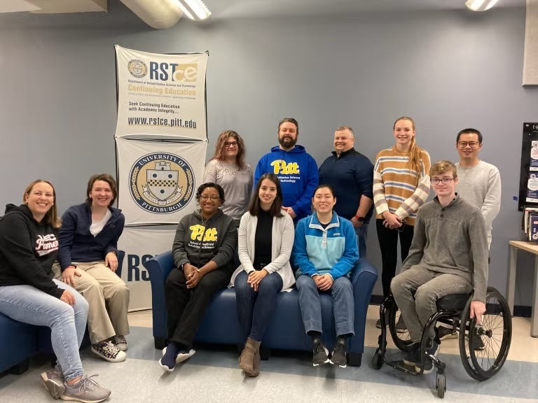 A group of people sitting and standing together near a sign that reads "Rehabilitation Science and Technology Continuing Education."