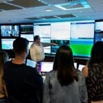 A man pointing at several screens displaying data, while four students gather around to listen.