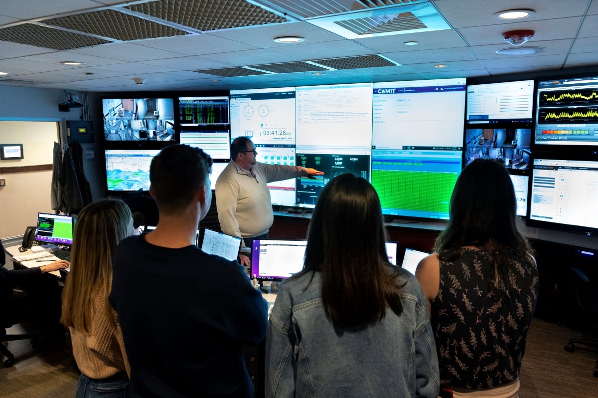 A man pointing at several screens displaying data, while four students gather around to listen.