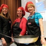 Three women wearing red chef hats and black chef aprons standing together over a large frying pan that is sitting on a table.