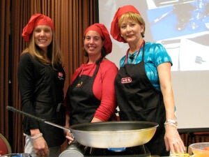 Three women wearing red chef hats and black chef aprons standing together over a large frying pan that is sitting on a table.