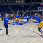 A woman with long brown hair wearing a blue Pitt Panthers shirt and black pants smiling on a basketball court while other players are running around her.