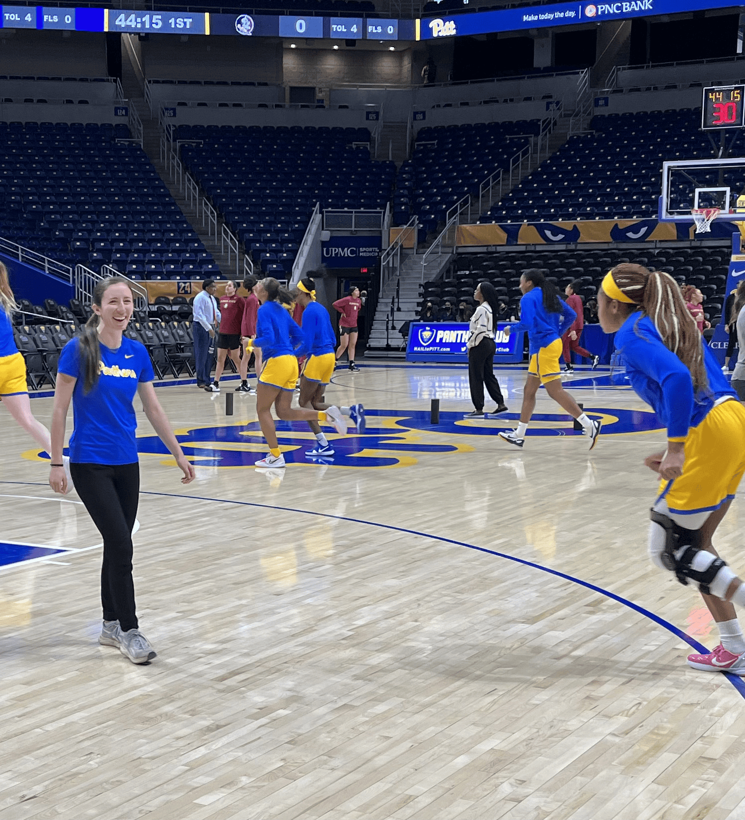 A woman with long brown hair wearing a blue Pitt Panthers shirt and black pants smiling on a basketball court while other players are running around her.