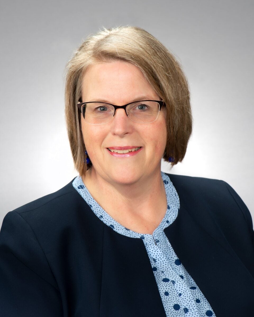 A woman with short blonde hair wearing a black blazer over a light blue blouse with large and small dark blue dots.