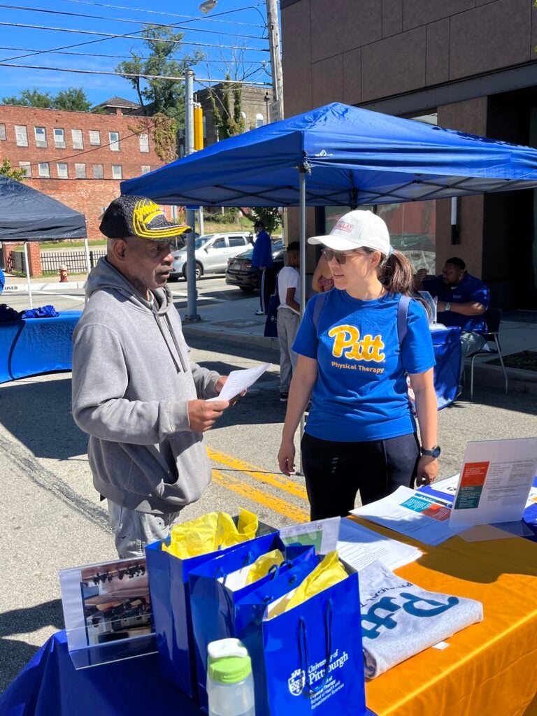 A man wearing a gray sweatshirt and a black and yellow hat standing next to a woman wearing a white hat with a blue Pitt t-shirt and black pants.