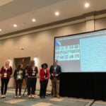 A group of men and women wearing lanyards standing together in front of a projected screen holding an award.