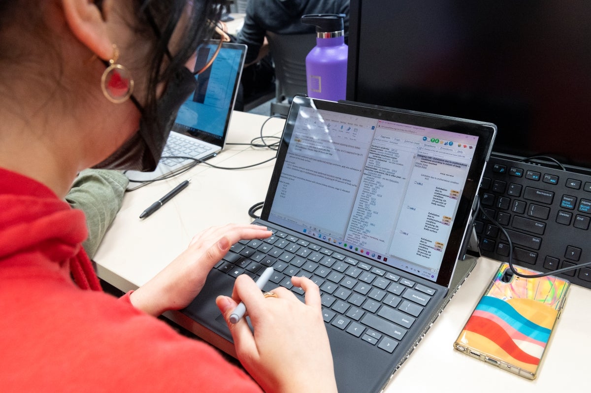 Woman with black hair wearing a face mask and a red sweatshirt looking at a laptop while holding a stylus pen.