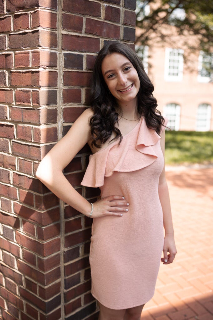 A woman with brown hair wearing a peach dress standing in front of a brick wall.