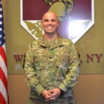 A bald man in an army camouflage uniform standing next to an America flag.