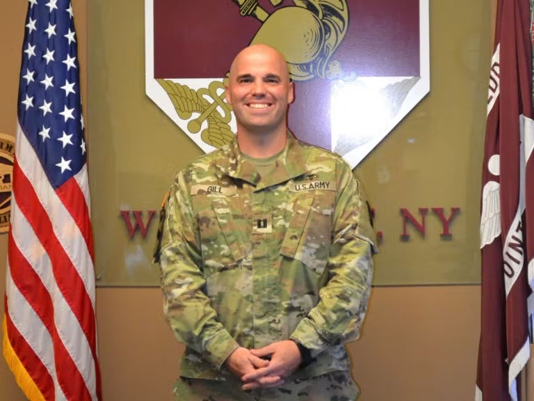 A bald man in an army camouflage uniform standing next to an America flag.